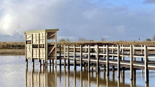 The wooden boardwalk leading to a small bird screen over shallow wetlands at Sandilands Nature Reserve, with reflections in calm water under a cloudy sky.